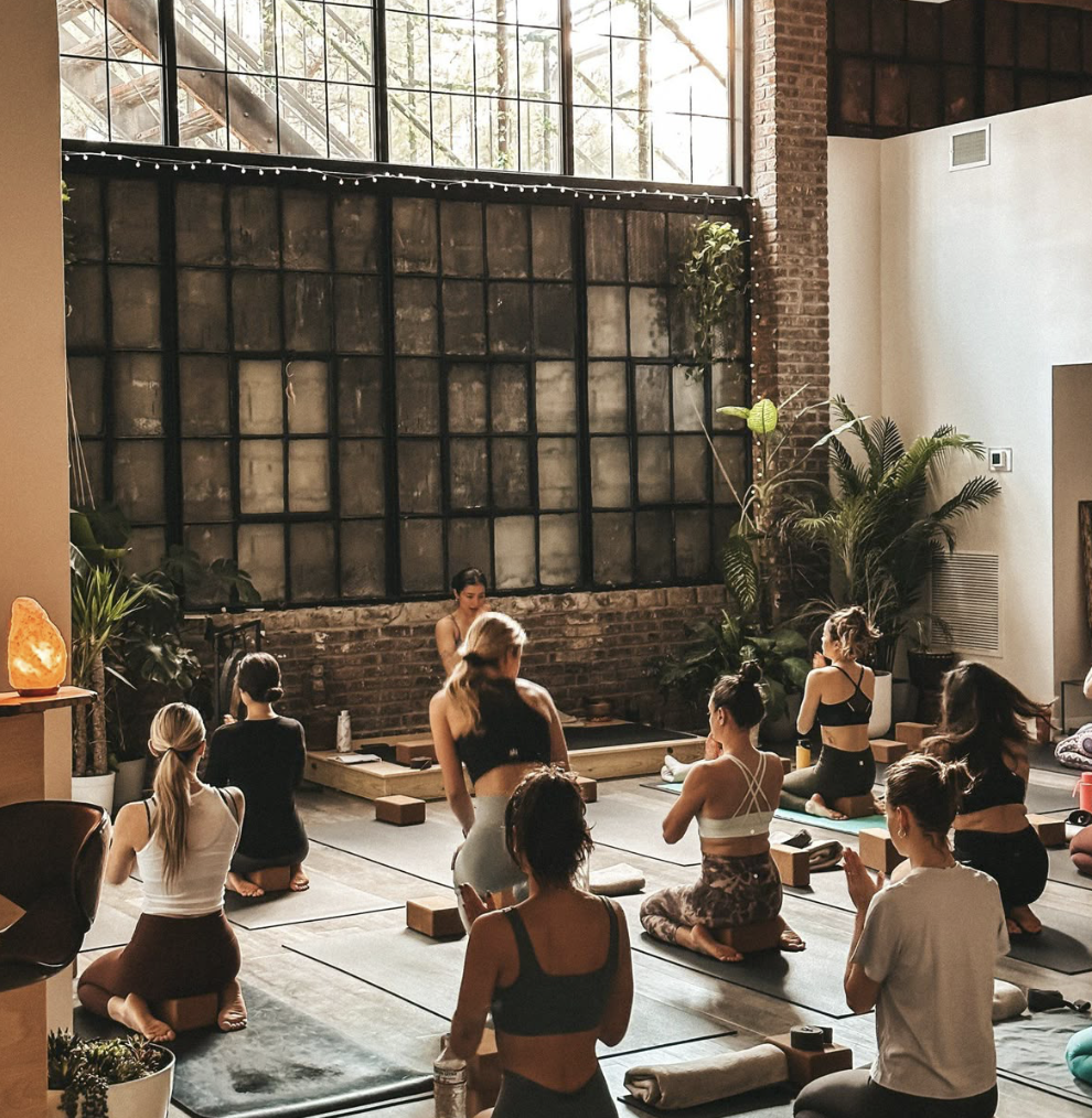 Women in a meditative yoga class in an industrial-chic studio with large windows, plants and a glowing salt lamp