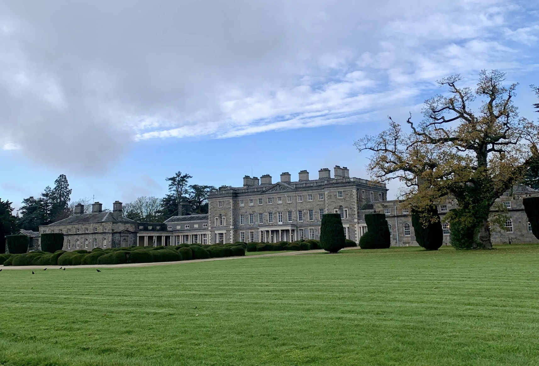 The grey stone facade of Carton House, a Georgian country house set behind a wide manicured lawn and low hedges under a blue sky