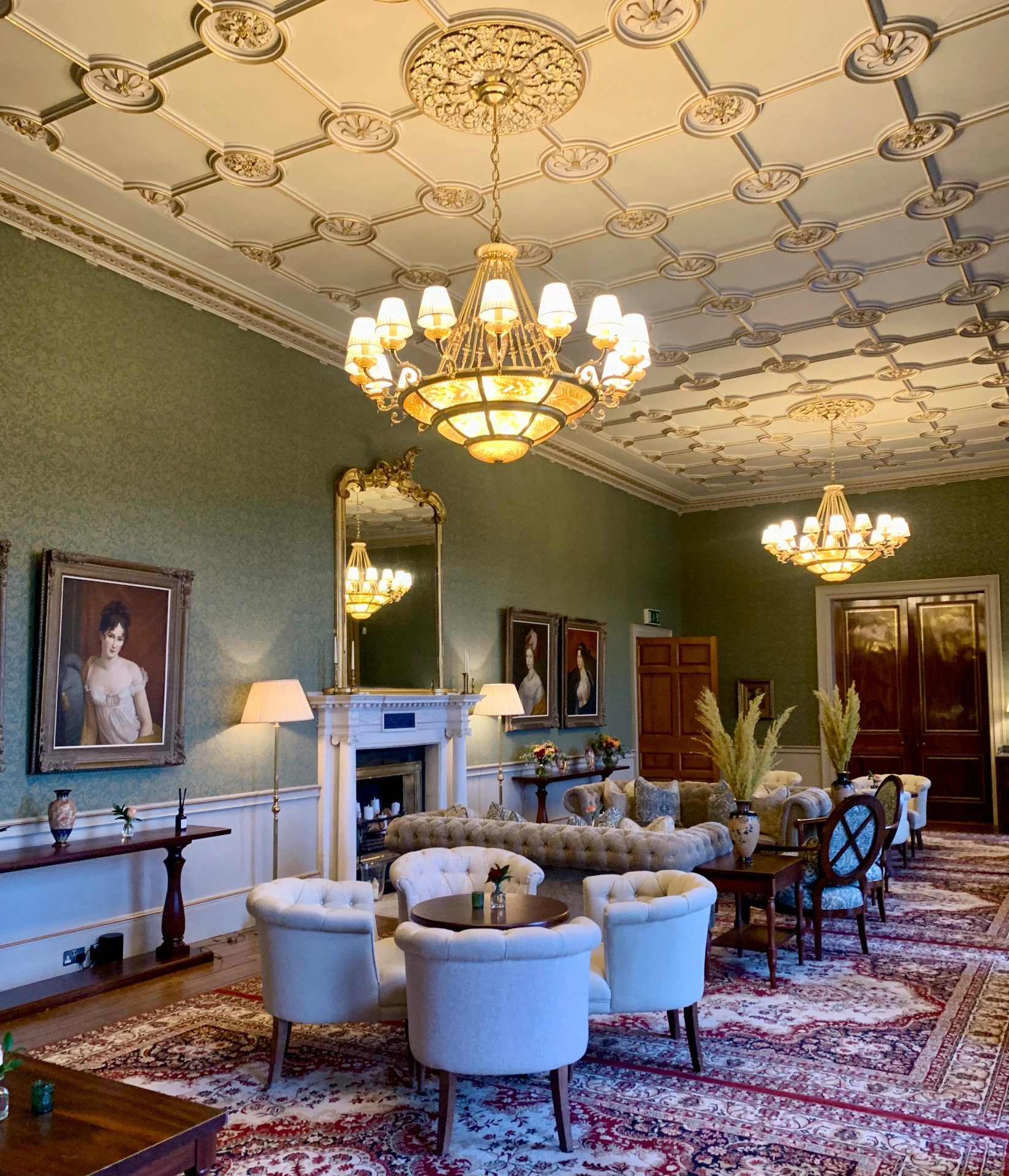 The drawing room at Carton House, with sage green walls, a marble fireplace, ornate chandeliers and tufted armchairs around a Persian rug